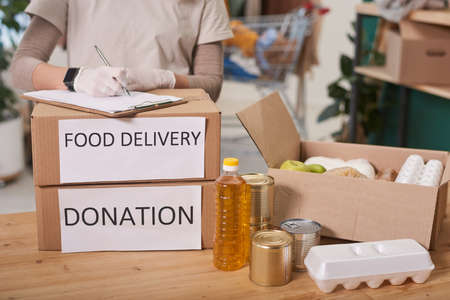 Unrecognizable volunteer standing at table filling in checklist of food supply boxes ready for donationの写真素材