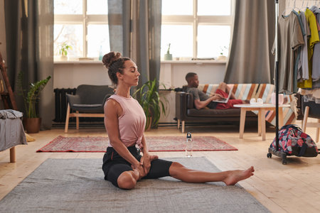 Horizontal portrait shot of modern sporty young woman sitting on floor stretching her leg while her...