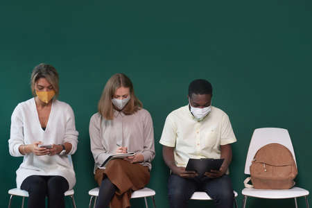 Two women and young man wearing protective masks sitting on chairs against tidewater green wall waiting for job interview in queueの写真素材