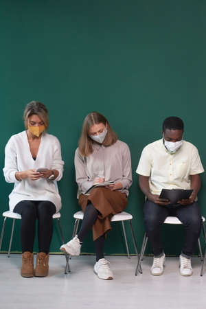 Conceptual shot of three modern people wearing protective masks sitting on chairs against tidewater green wall waiting for job interviewの写真素材