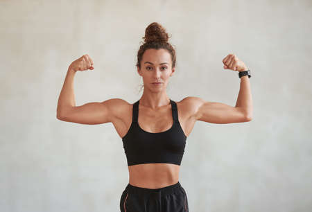 Portrait of modern young adult Caucasian woman wearing black sports outfit demonstrating her biceps on cameraの写真素材