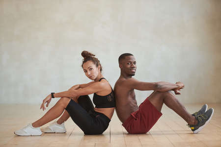 Studio shot of sporty young Black man and Caucasian woman sitting back to back in loft studio room looking at cameraの写真素材