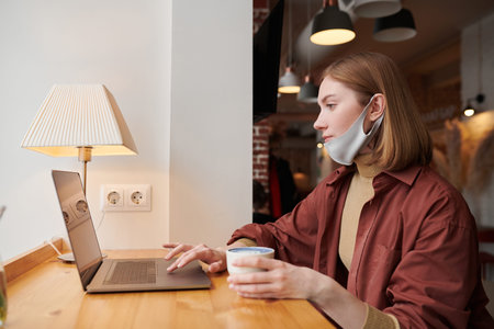Medium portrait of young Caucasian woman wearing mask having coffee break in modern cafe sitting at...