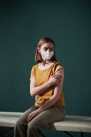 Conceptual portrait of young woman wearing protective mask sitting against tidewater green wall background pressing cotton pad after getting vaccineの写真素材