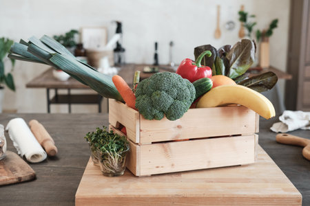 Wooden box full of various vegetables and fruits placed on board with growing plants into glasses in modern kitchenの写真素材