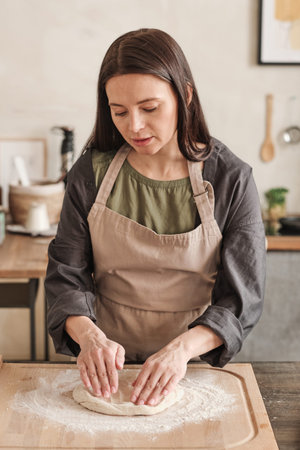 Serious young woman in apron standing at table with wooden board and kneading dough while cooking...