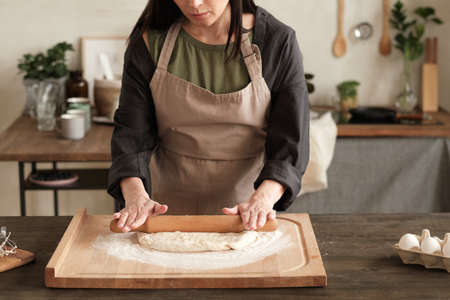 Busy woman in apron standing at table and rolling dough with pin on wooden board in modern kitchenの写真素材