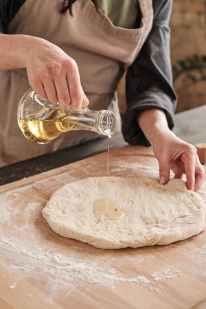 Close-up of female hand pouring oil from jug onto dough while making homemade pizzaの写真素材