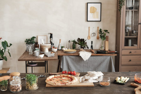 Prepared pizza on wooden board placed on table with ingredients and jars of spices, table with...