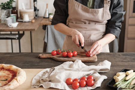 Close-up of unrecognizable woman in apron standing at counter with vegetables and cutting tomatoes...
