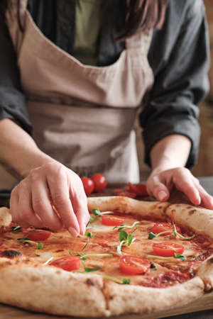 Close-up of unrecognizable woman in apron putting pea leaves on pizza with tomatoes while preparing it for familyの写真素材