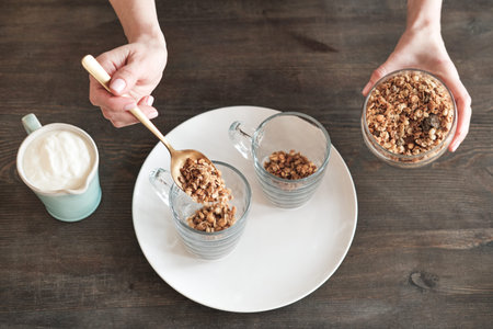 Directly above view of unrecognizable woman putting muesli into mug while mixing it with yogurt for...