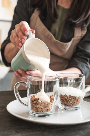 Close-up of unrecognizable woman in apron pouring yogurt into mug with muesli while making healthy...