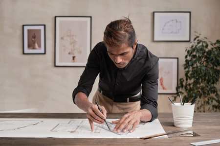 Concentrated young Indian building engineer in black shirt leaning on table and using ruler while...