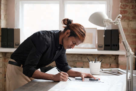 Serious middle-aged Black designer with red hair standing at wooden desk and editing sketch with pencil while working on blueprintの写真素材