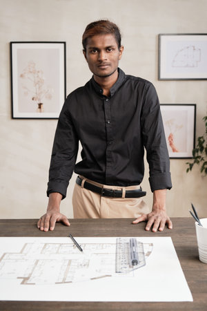 Portrait of content young Indian architect in black shirt standing at wooden table with blueprint,...