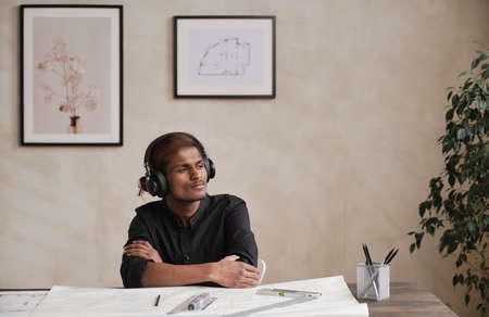 Pensive dreamy inspired Indian architect in wireless headphones sitting with crossed arms at table with blueprint and looking aside in officeの写真素材