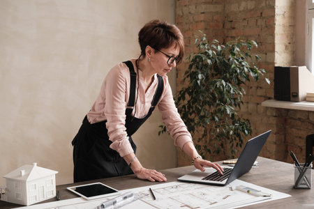 Concentrated female architectural designer in eyeglasses standing at desk with blueprint and editing...