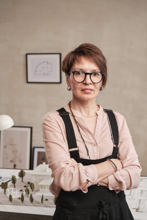 Portrait of smiling confident mature architect in eyeglasses standing with crossed arms against...