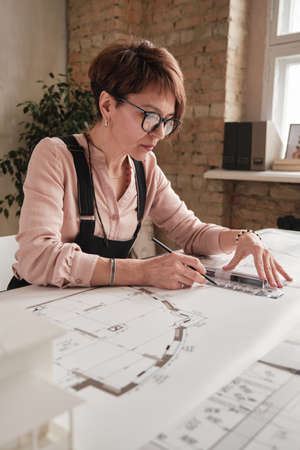 Serious skilled mature draftswoman in eyeglasses sitting at table and working with sketch using ruler and pencil in officeの写真素材