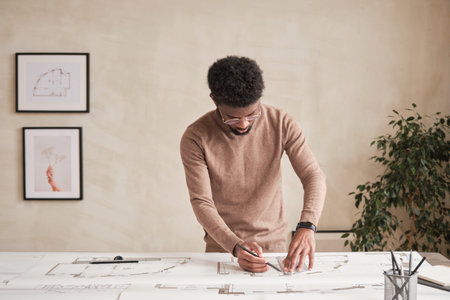 Serious young African-American architect in glasses standing at desk and drafting blueprint while...