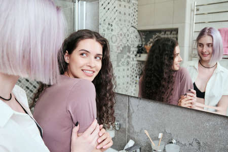 Purple-haired woman touching shoulder of girlfriend in bathroom while they doing morning routine togetherの写真素材