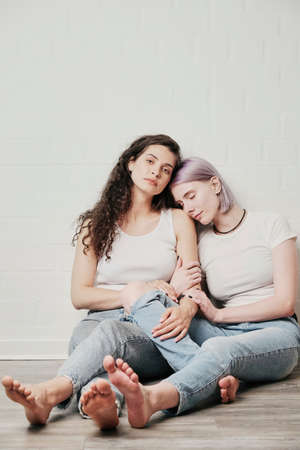 Relaxed purple-haired woman sitting on floor and sleeping on shoulder of girlfriend while feeling comfort and calmnessの写真素材