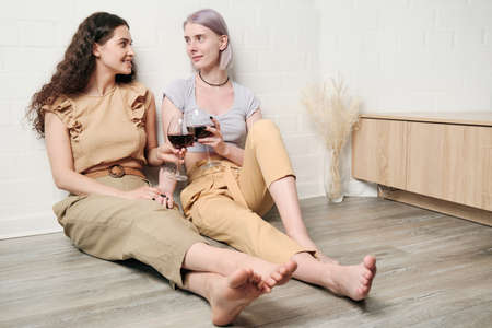 Happy young Caucasian lesbians sitting on floor and clinking wineglasses while drinking wine at homeの写真素材