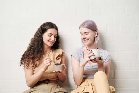 Smiling Caucasian woman sitting against wall and feeding female lover with spoon during breakfastの写真素材