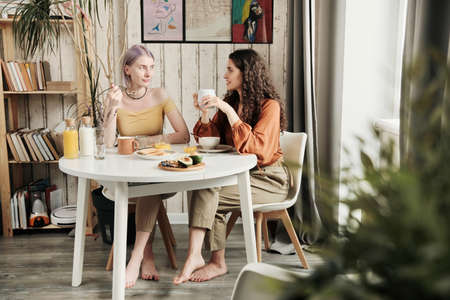 Attractive young lesbians sitting at dining table and drinking coffee while chatting during breakfast at homeの写真素材