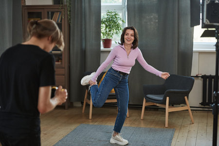 Operator shooting cheerful woman standing on one leg in living room while making video for social...