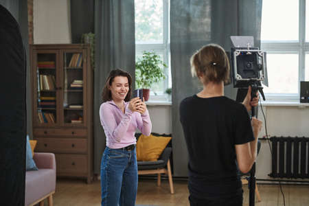 Rear view of assistant adjusting strobe light in living room while blogger shooting video on smartphoneの写真素材