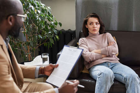 Depressed young Caucasian woman sitting with crossed arms on sofa and listening to psychiatristの写真素材
