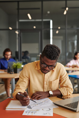 Busy young African-American businessman in eyeglasses sitting in shared workspace and analyzing...
