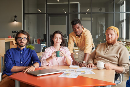 Group photo of confident young multi-ethnic colleagues sitting at table with papers and drinking...