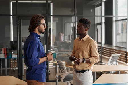 Young multi-ethnic managers standing in coworking space and discussing new project while one of them...