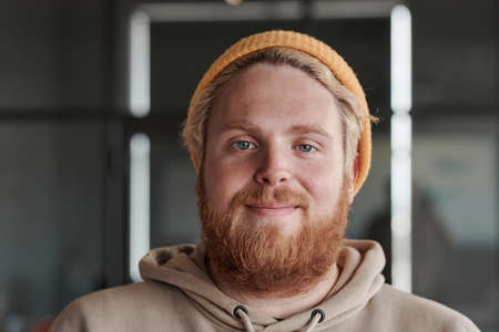 Headshot of satisfied young red-bearded Caucasian man in bright hat posing in modern officeの写真素材