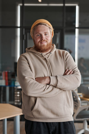 Portrait of smiling young bearded Caucasian man in hat and hoodie standing with crossed arms in...