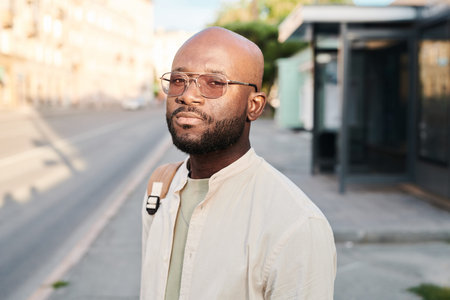 Portrait of serious young black man in sunglasses standing at bus station and waiting for transport...