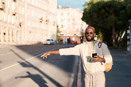 Handsome young Black man in earphones sticking out arm while catching taxi in city using smartphone appの写真素材