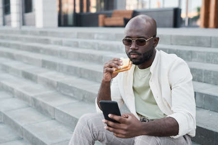 Serious young African-American city man in sunglasses sitting on stairs and eating sandwich while using smartphone outdoorsの写真素材