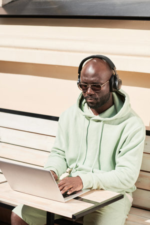 Serious young African-American man in headphones and sunglasses sitting at table and using laptop...