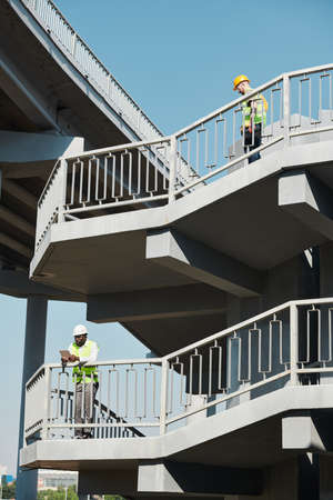 Serious busy men in hardhats and green vests working at construction site of apartment building and discussing infrastructure while contemplating cityの写真素材