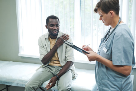 African American patient sitting on examination couch and complaining on shoulder pain to doctor in...