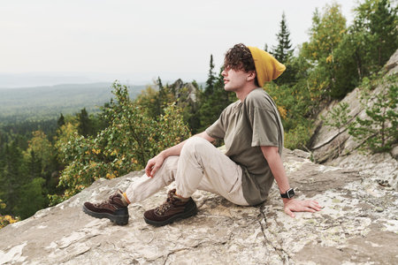 Dreamy young Caucasian hiker in yellow hat and boots sitting on big stone and contemplaing forest landscapeの写真素材