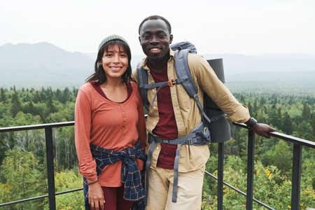Happy multi-ethnic couple standing at railings against forest landscape and looking at camera at hikeの写真素材