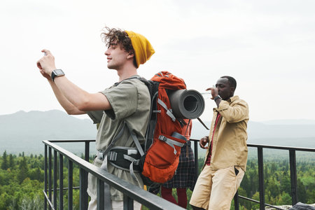 Smiling young hiker with backpack standing on bridge and saving moments while photographing...
