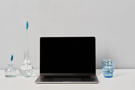 Office workplace with laptop, vases with decorative flowers and glass of water in white and blue...