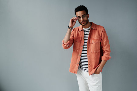 Portrait of content stylish young African American man in coral shirt holding hand in jeans pocket...