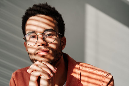 Headshot of content young black man with stubble leaning head on clasped hands in shadow of blinds